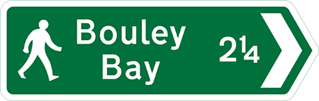 Green road sign with image of person walking and white text showing destination, distance and direction